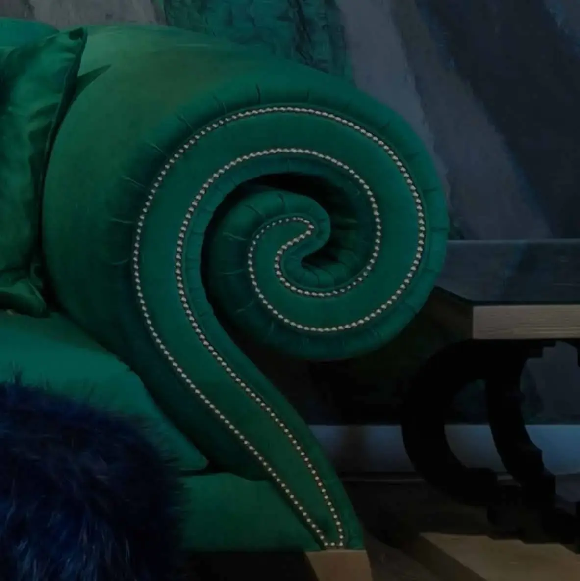 Detail of an emerald velvet sofa arm curled into a spiral, its edge traced with studded piping