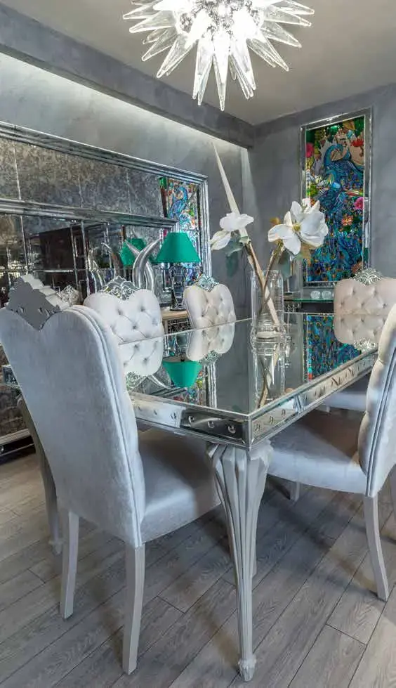 Mirror-based dining table with buttoned cream chairs, a teal table lamp and a peacock panel on the wall behind
