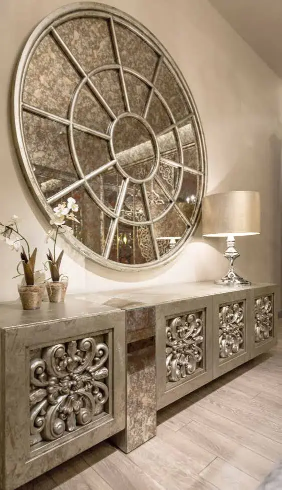 Large round window-pane mirror hung above a silver-leaf console with scrolled relief panels and potted orchids