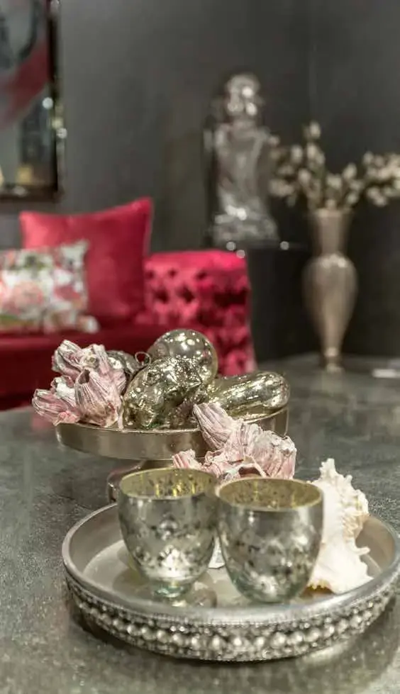 Silver cups and pinecone ornaments arranged on a beaded tray, with a crimson sofa behind